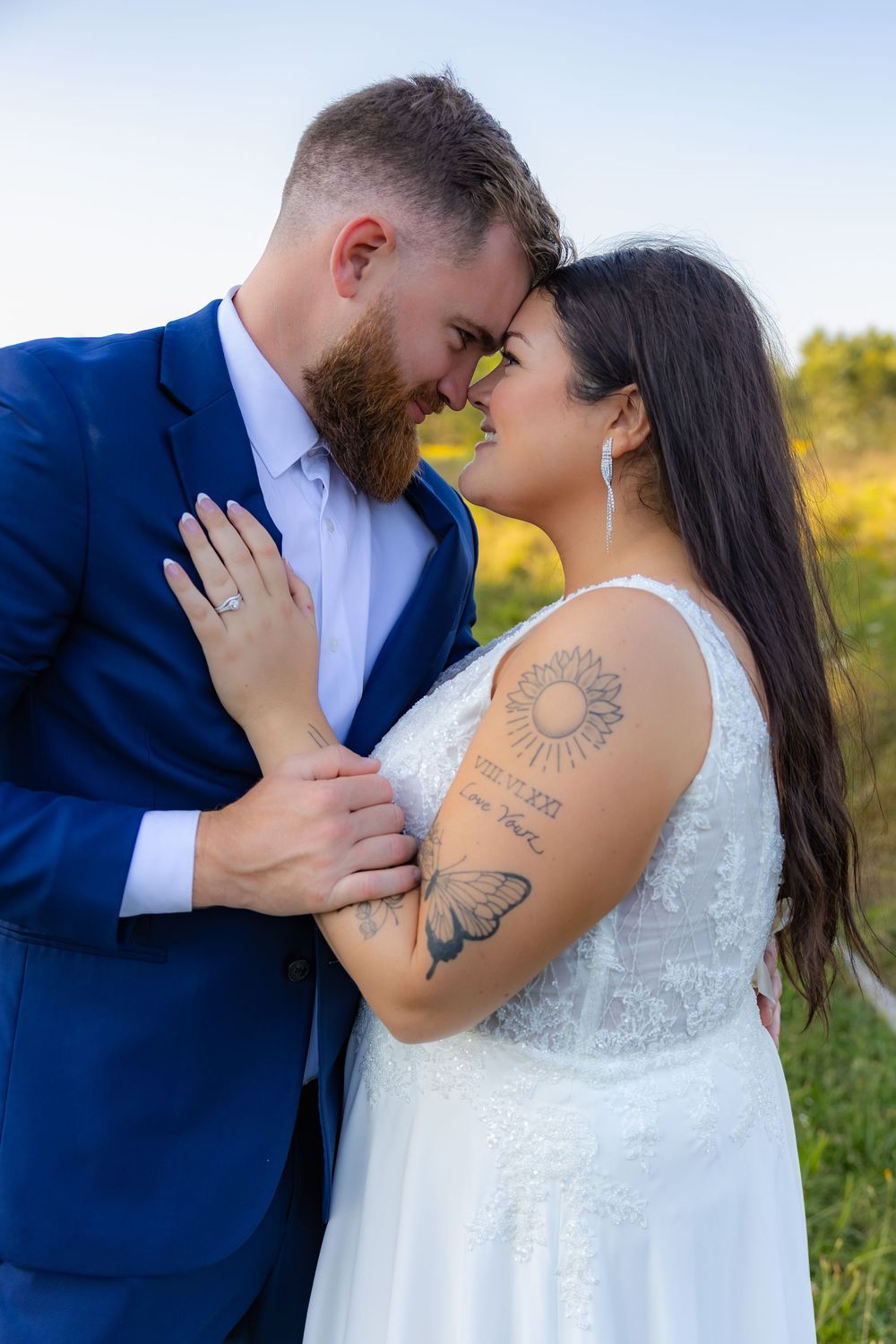 A couple shares an intimate moment in a field wearing wedding attire with visible tattoos on display.