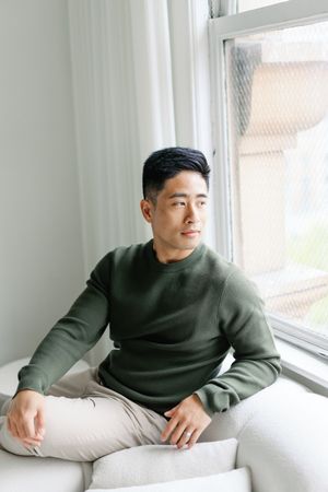 A person wearing a dark turtleneck sweater lounges on a white couch near a sunny window in a minimalist setting.