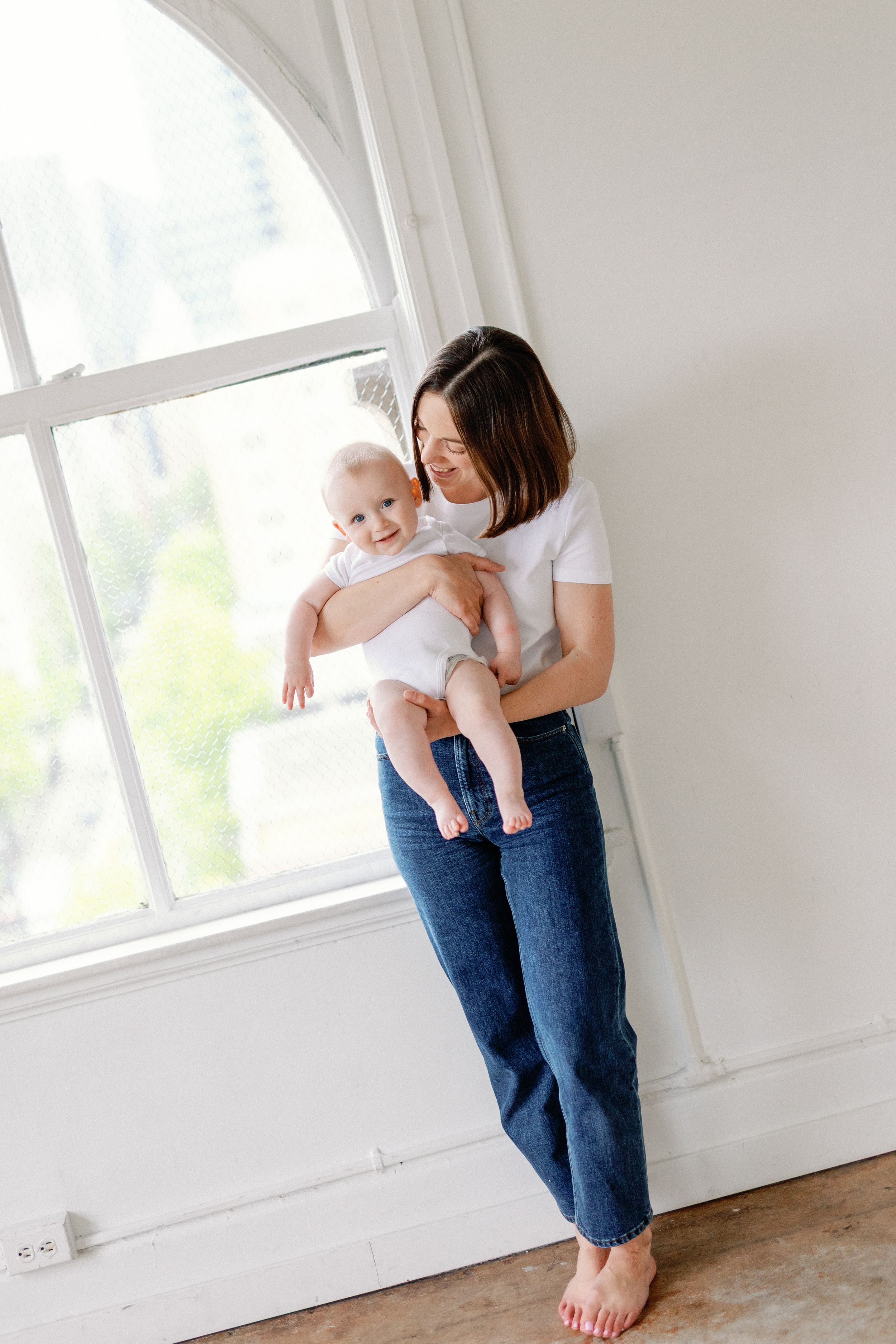 A parent wearing blue jeans and white t-shirt holds a baby by a bright window in a minimalist white room.