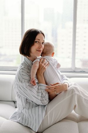A mother in a striped shirt cradles and nurtures her baby in a bright, minimalist white room setting.