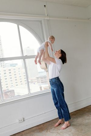 Parent in denim pants holds and lifts baby against white industrial space with large windows and city view.