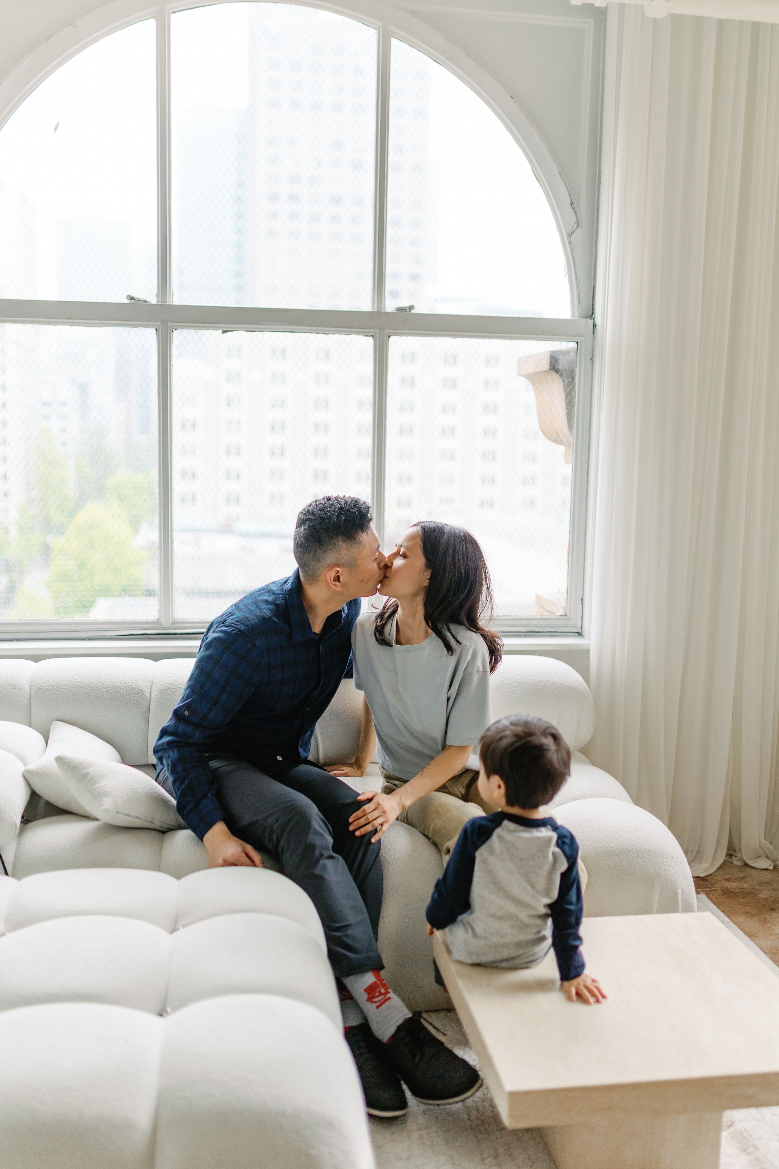 A family relaxes together on a white couch in a bright, modern living room with large windows.