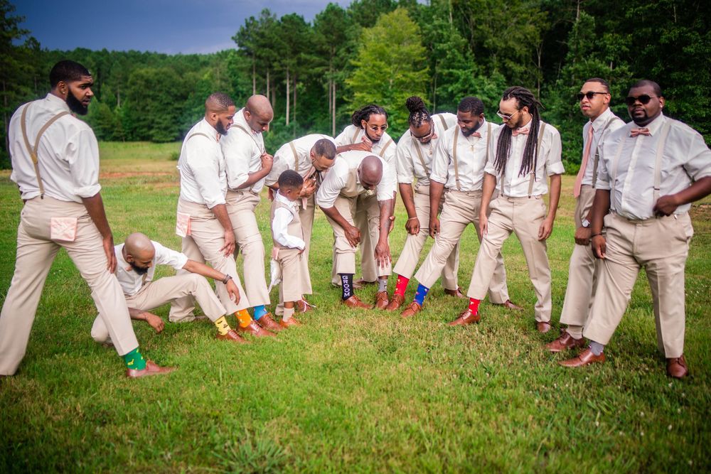 Groomsmen in beige suits pose playfully on grass during outdoor wedding.