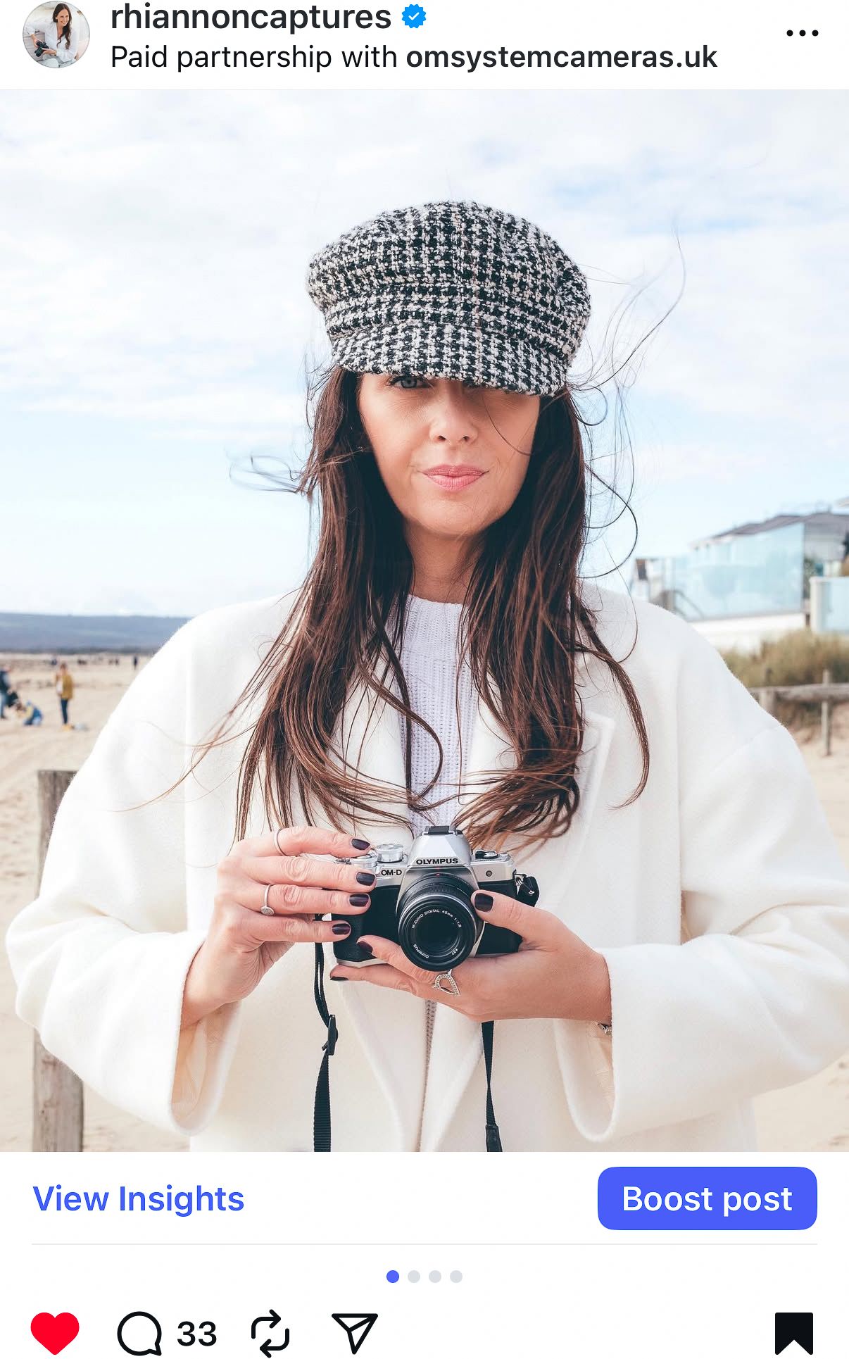 Individual in white attire holding a camera outdoors near a beach, wearing a checkered cap and demonstrating photography equipment.