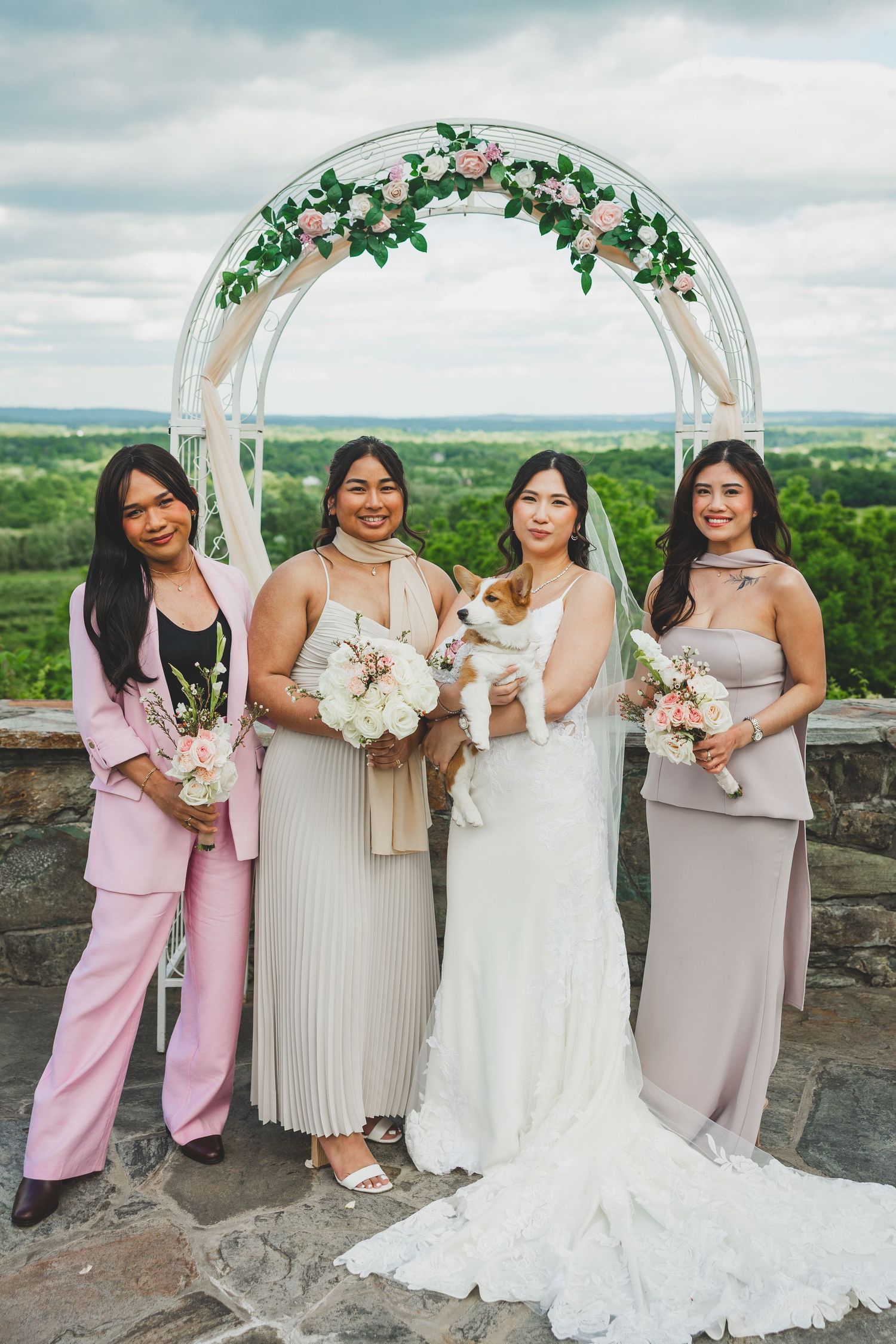 Four bridesmaids in pink and beige dresses holding bouquets pose together at an outdoor wedding venue.
