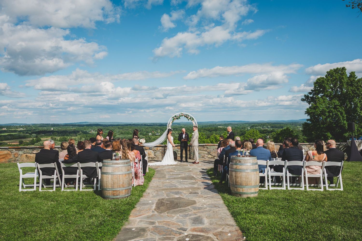 A panoramic view of an outdoor wedding ceremony setup on a hilltop with white chairs and wine barrels under blue skies.