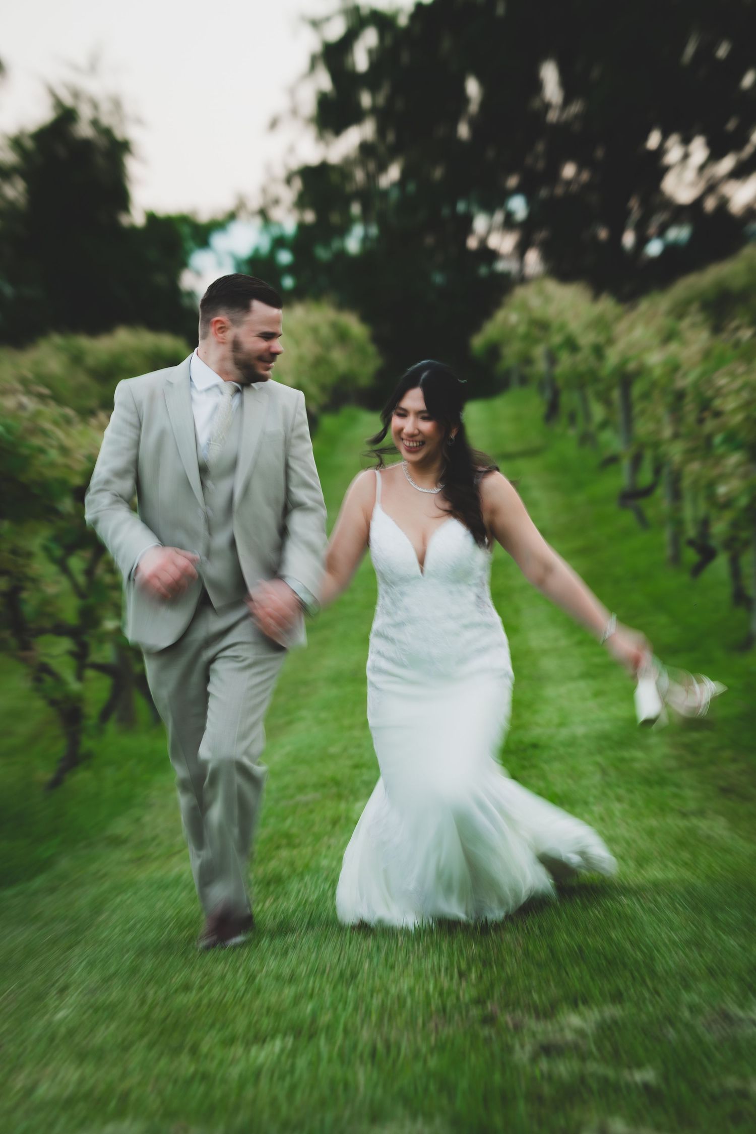 A couple in wedding attire runs playfully through a green apple orchard on their special day.
