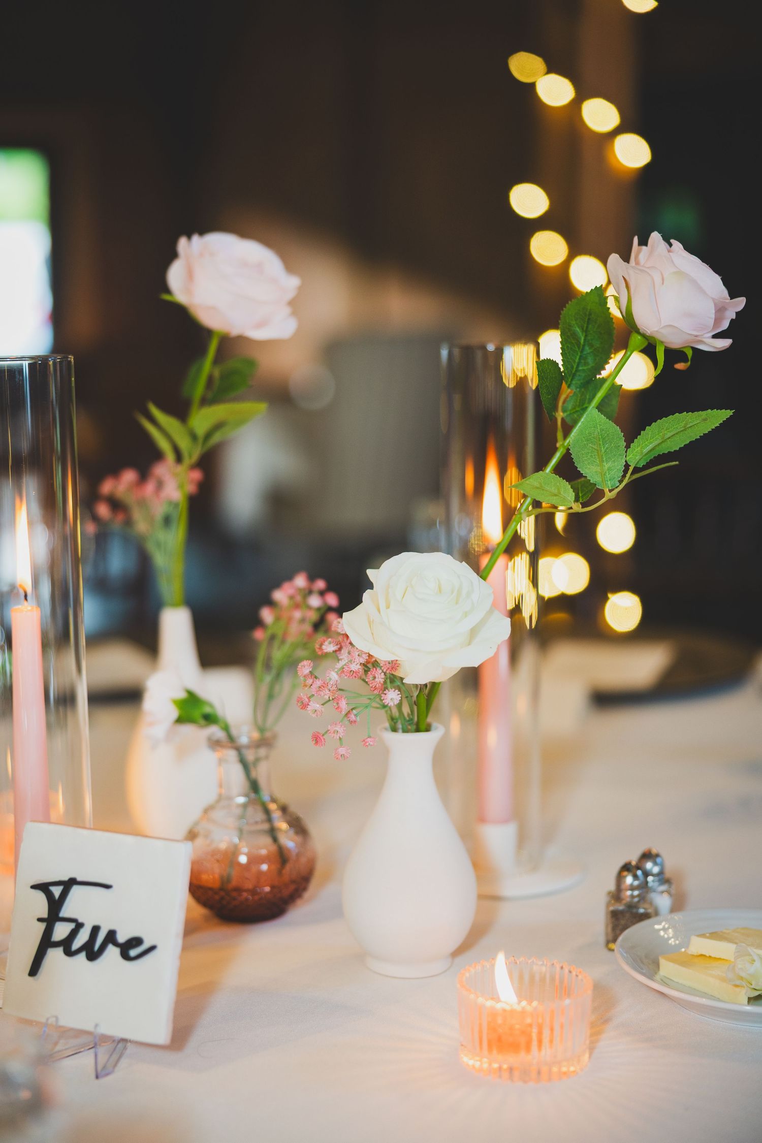 Elegant table setting with white roses in vase, pink candle holder and delicate string lights glowing in background.