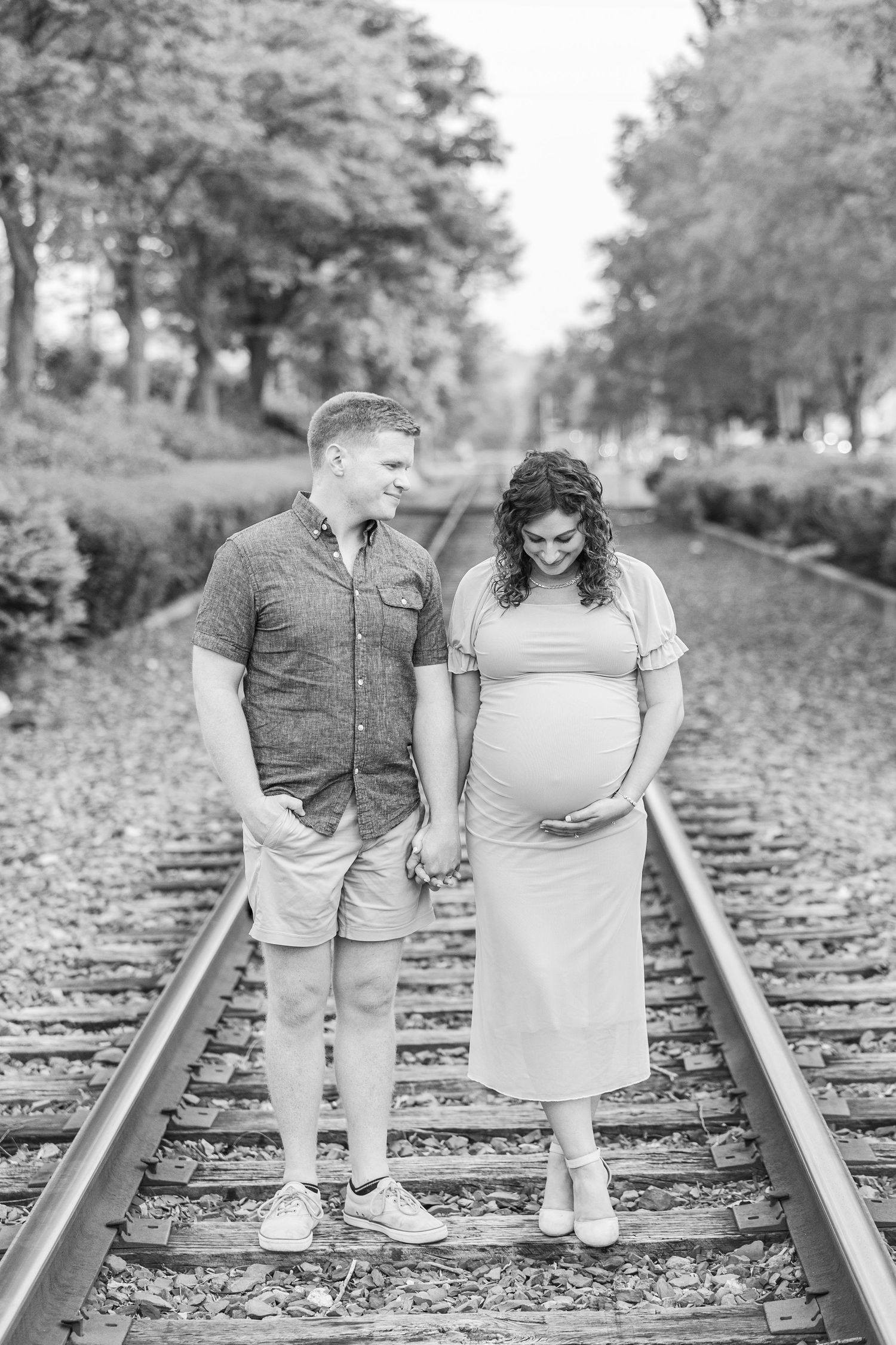 A couple poses together on railroad tracks surrounded by trees in black and white.