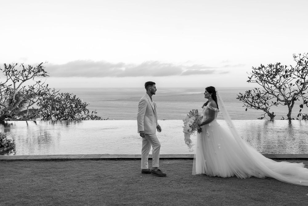 A black and white wedding photograph showing a couple standing by an infinity pool at sunset with ocean views.