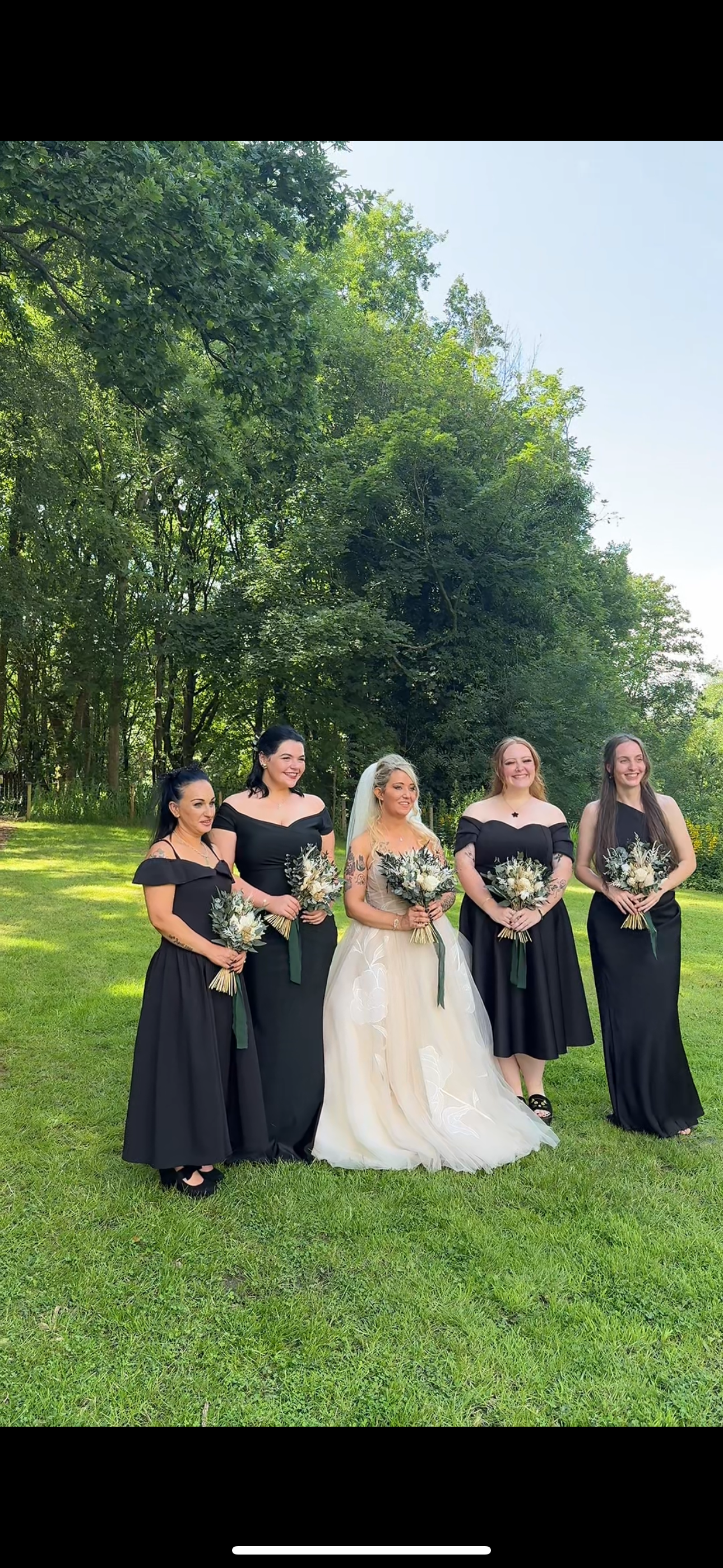 Bridal party in black dresses poses on grassy hill during summer wedding.