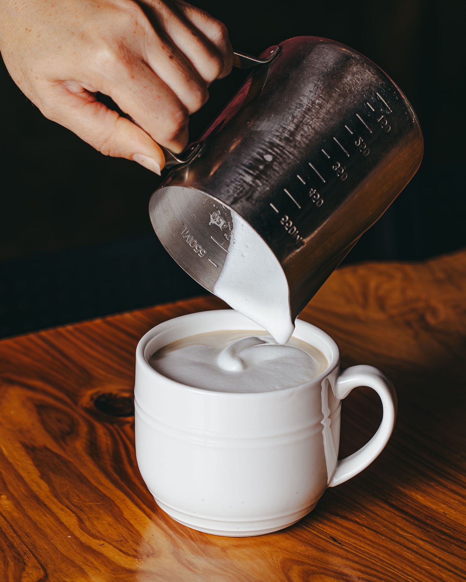 Steamed milk being poured from a glass measuring cup into a white coffee cup on a wooden table.