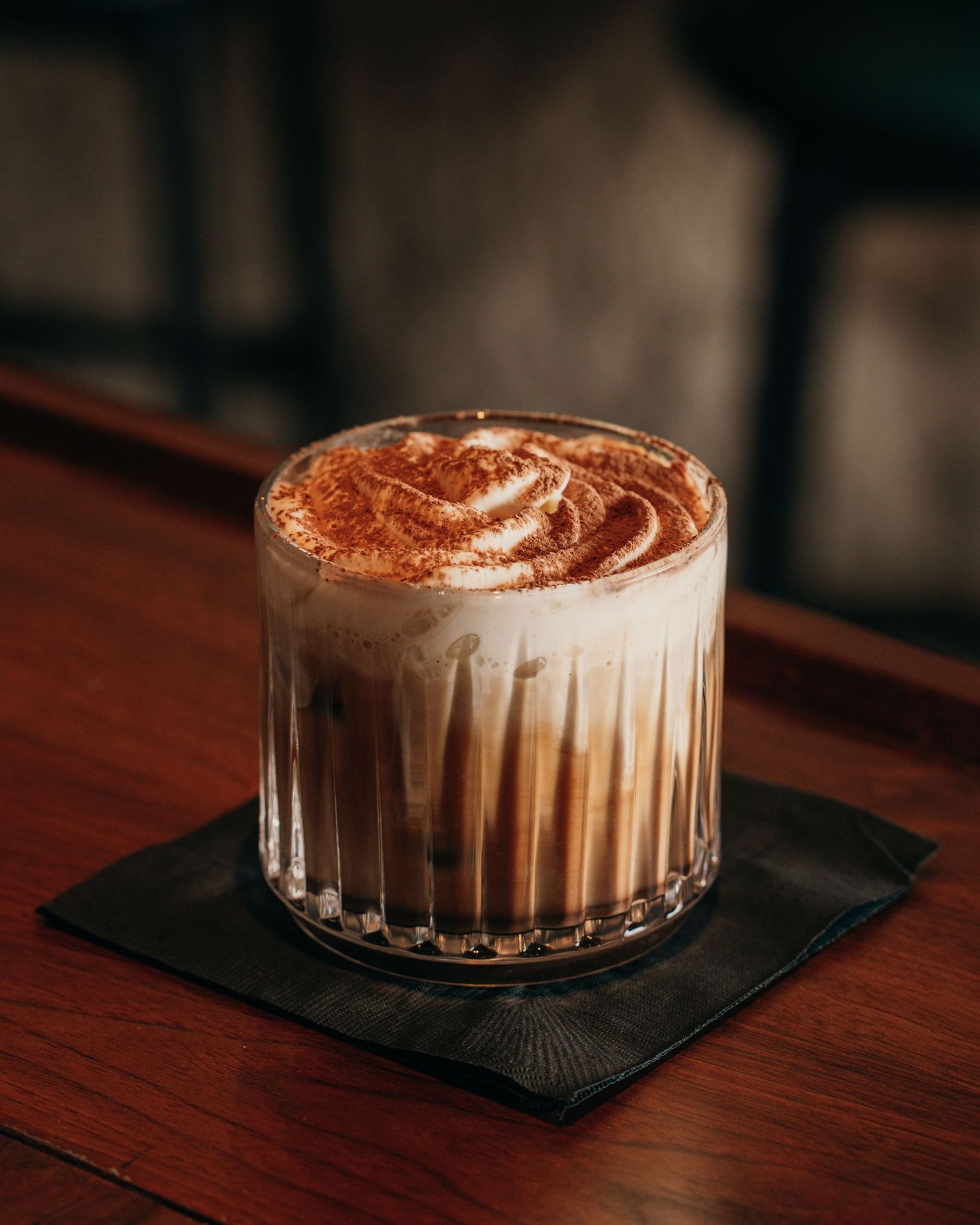A layered coffee drink with cream and cocoa powder served in a clear curved glass on a dark napkin.