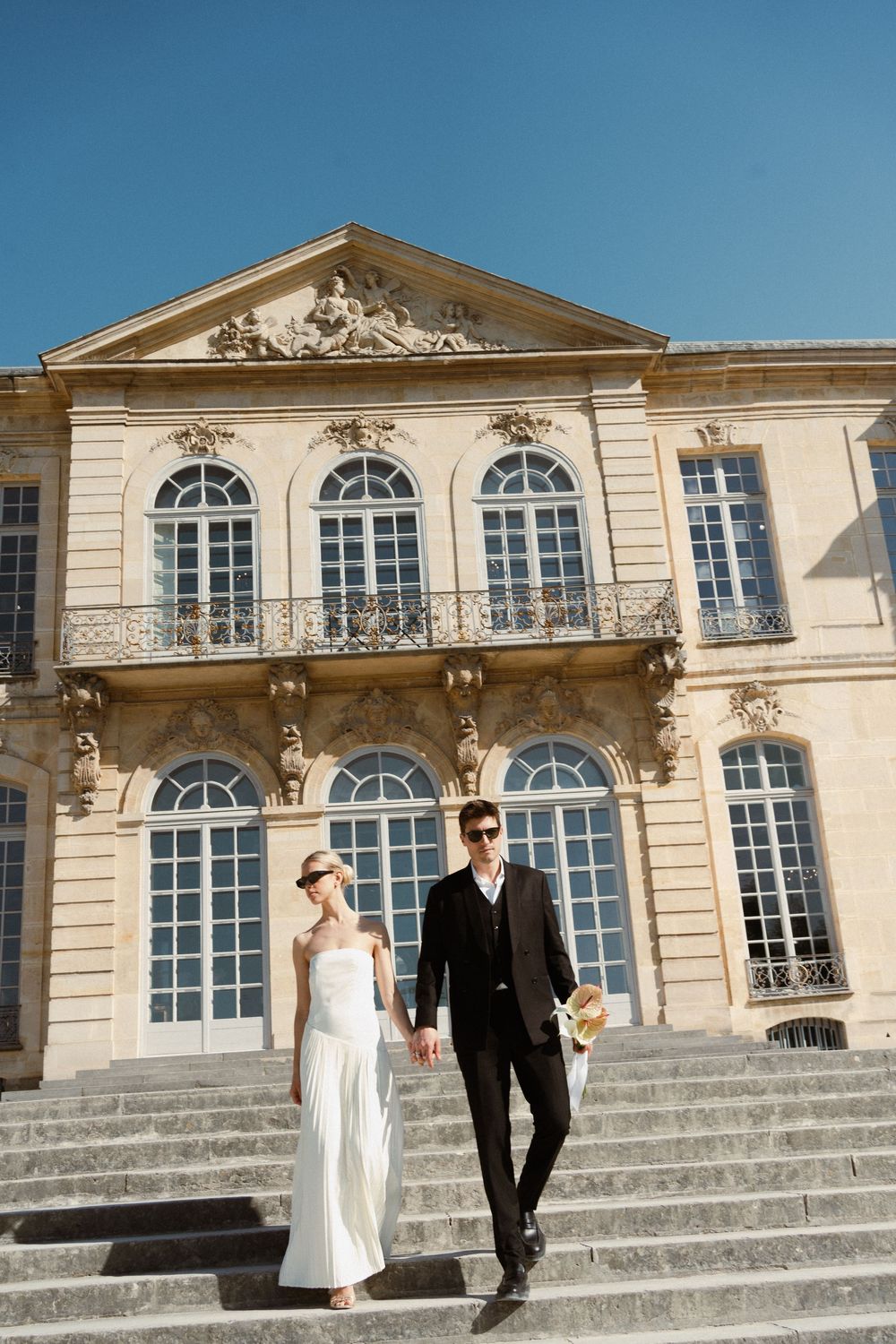 A couple walks down stone steps in front of an elegant historic manor house with arched windows and neoclassical architecture.