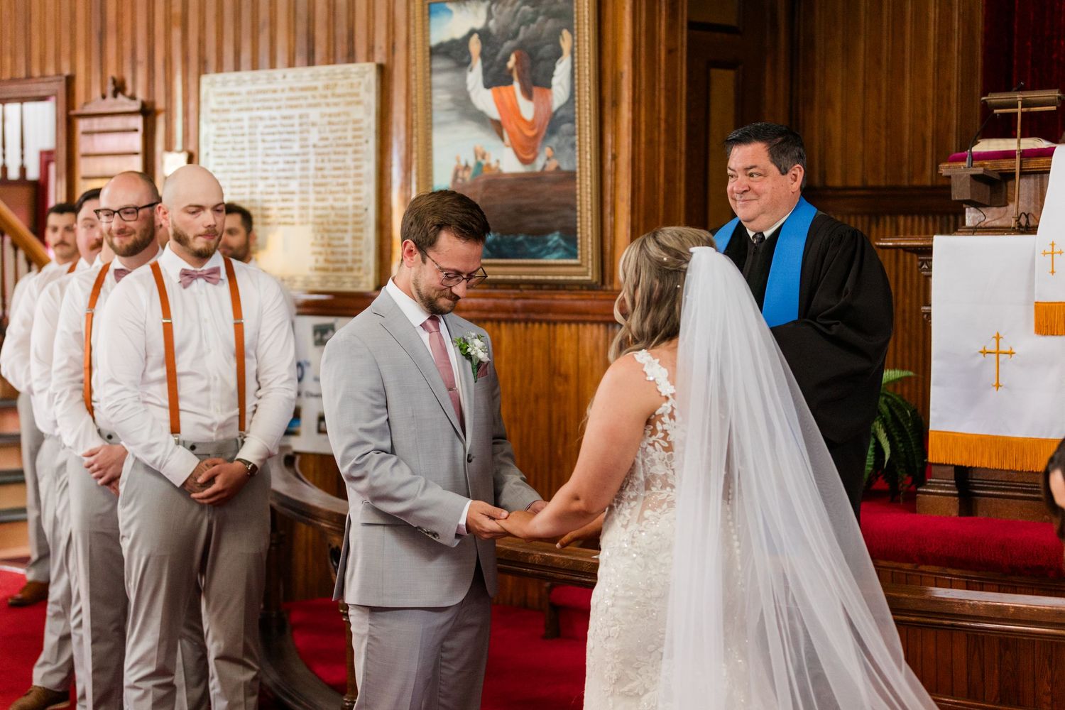 Bride in white lace dress shares touching moment during church ceremony with wedding party looking on.