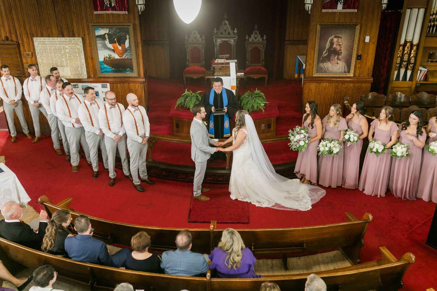 Wedding party poses for formal photos in a church sanctuary with red carpeting and wooden pews.
