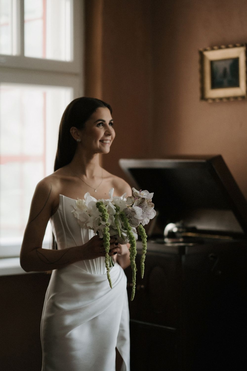 Bride in sleek white dress holds white flowers against warm-toned interior with window light.