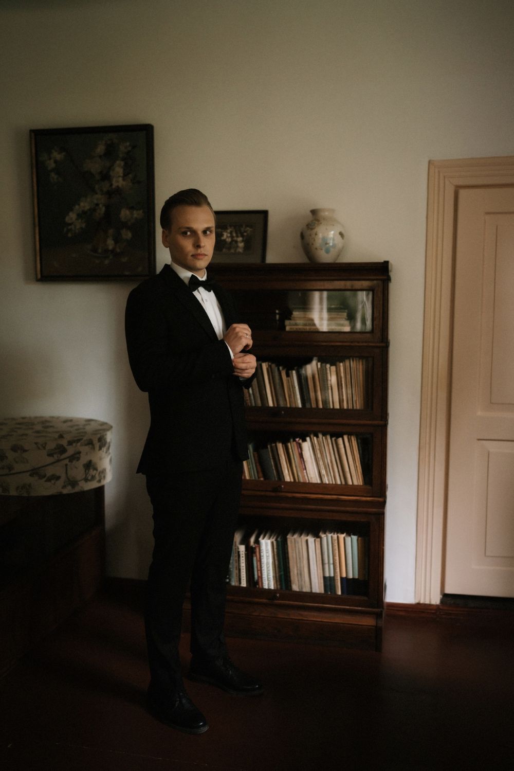 Moody interior shots showing a groom in a tuxedo standing in a room with bookshelves.