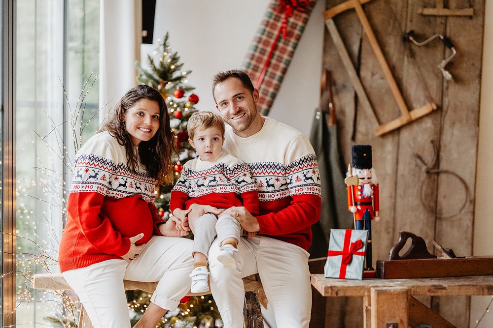 Family in matching Christmas sweaters posing with child near decorated tree and holiday decor.
