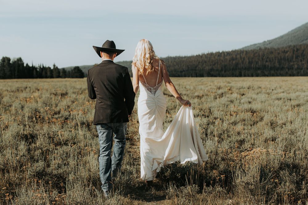 A couple walks hand in hand through a golden meadow with mountains and evergreen forest in the background.
