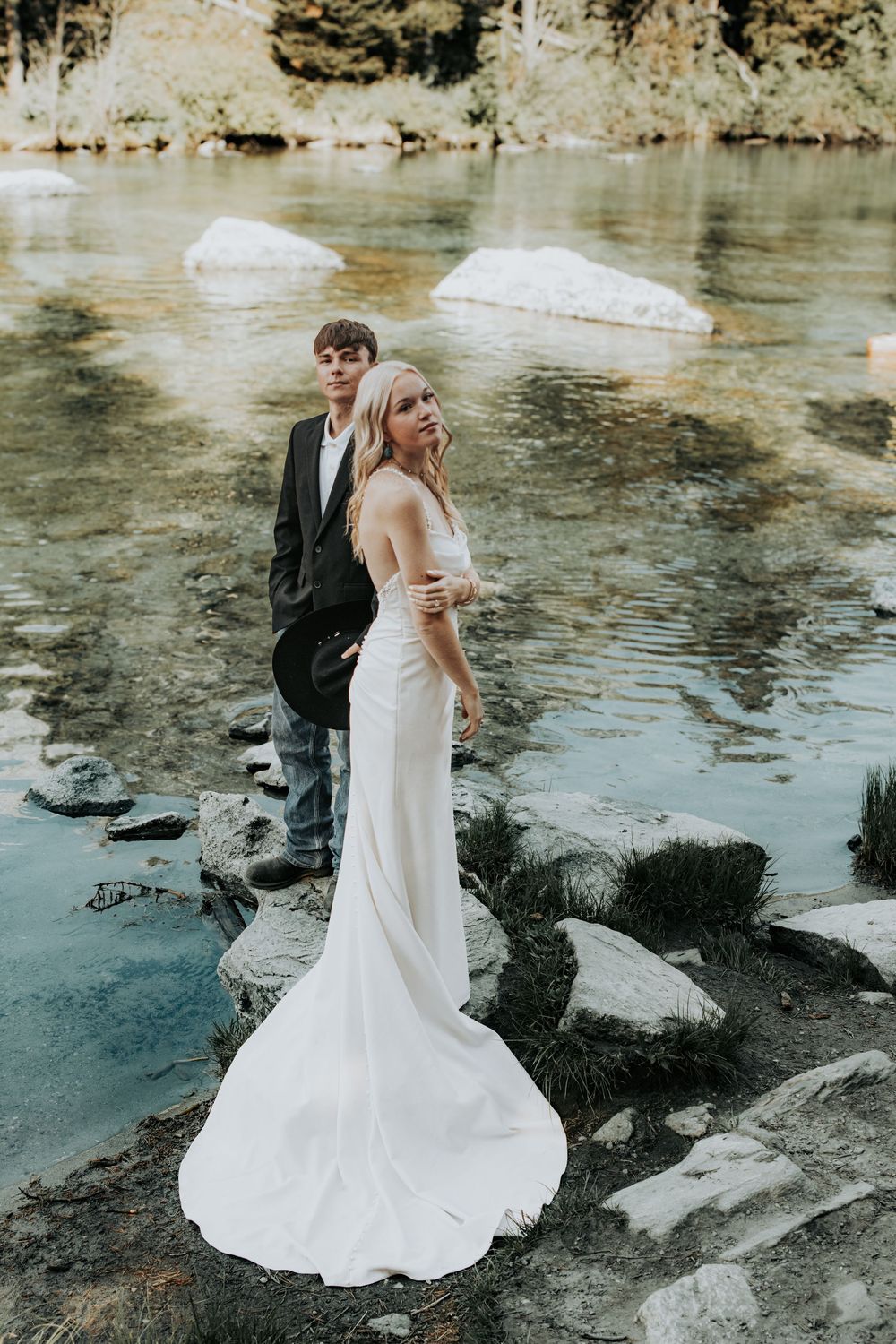 A couple in wedding attire stands on rocks by a serene lake during a nature-inspired outdoor photoshoot.