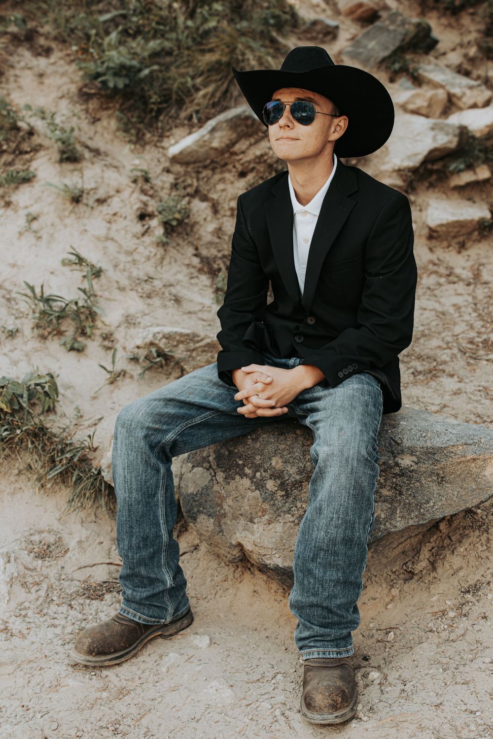 Stylish western-inspired portrait of someone wearing a black cowboy hat, blazer and jeans sitting on rocky terrain.