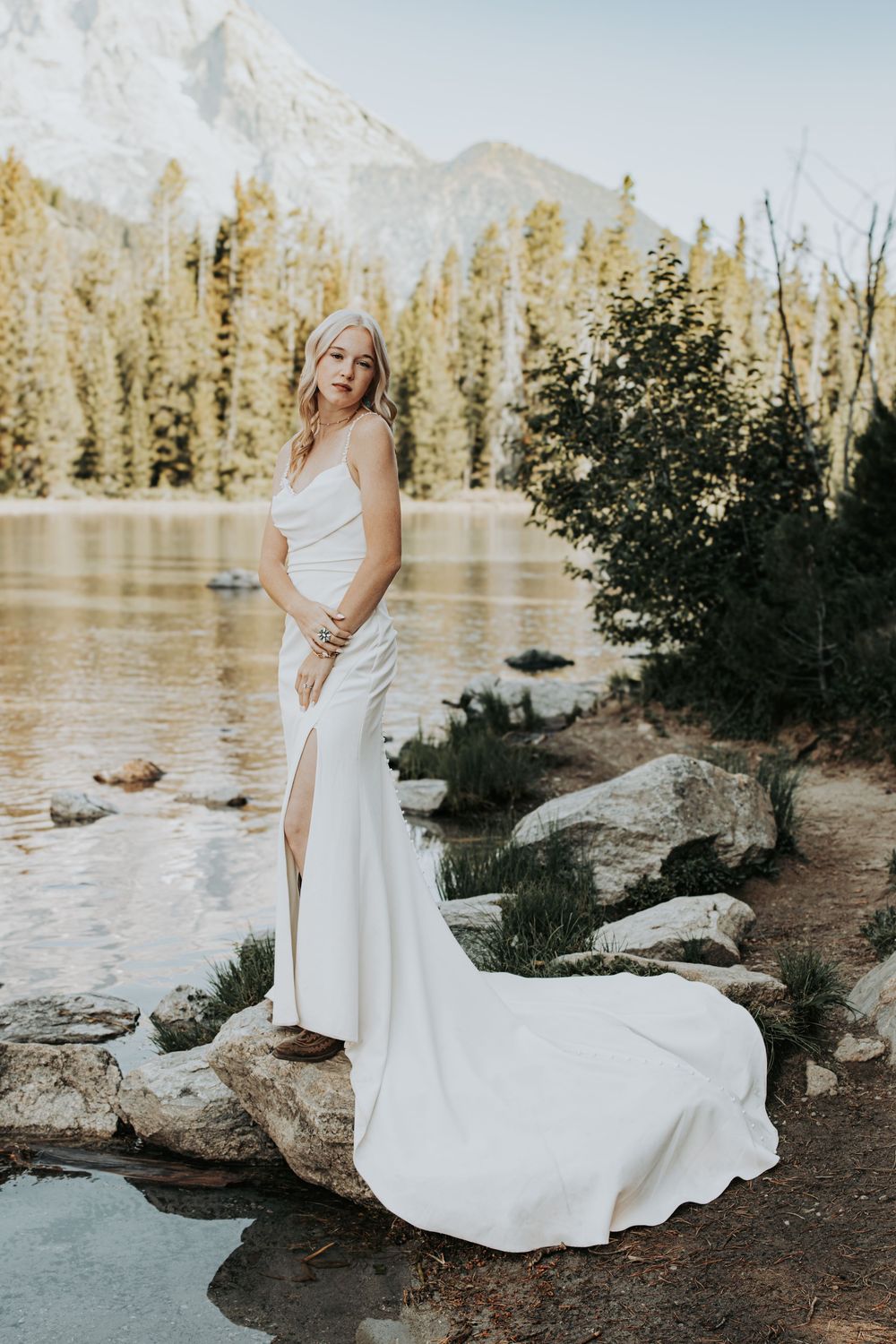 Wedding photography series by a mountain lake featuring a bride in a flowing white dress posing on rocky shore.