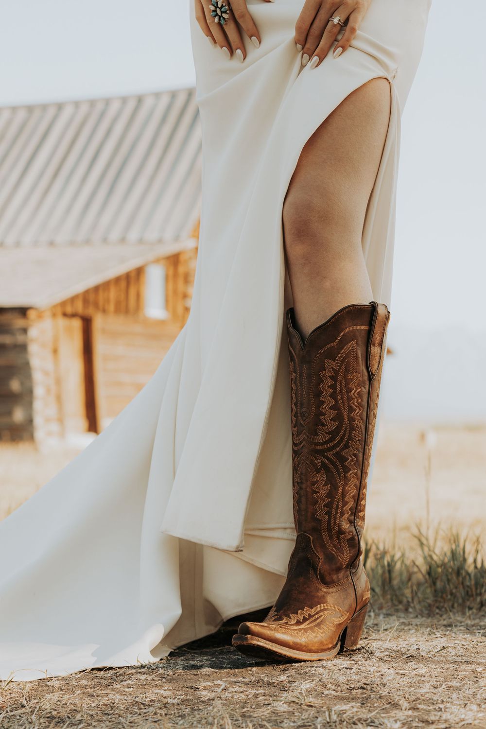 Detailed brown leather cowboy boots paired with flowing white wedding dress hem at rustic outdoor setting.