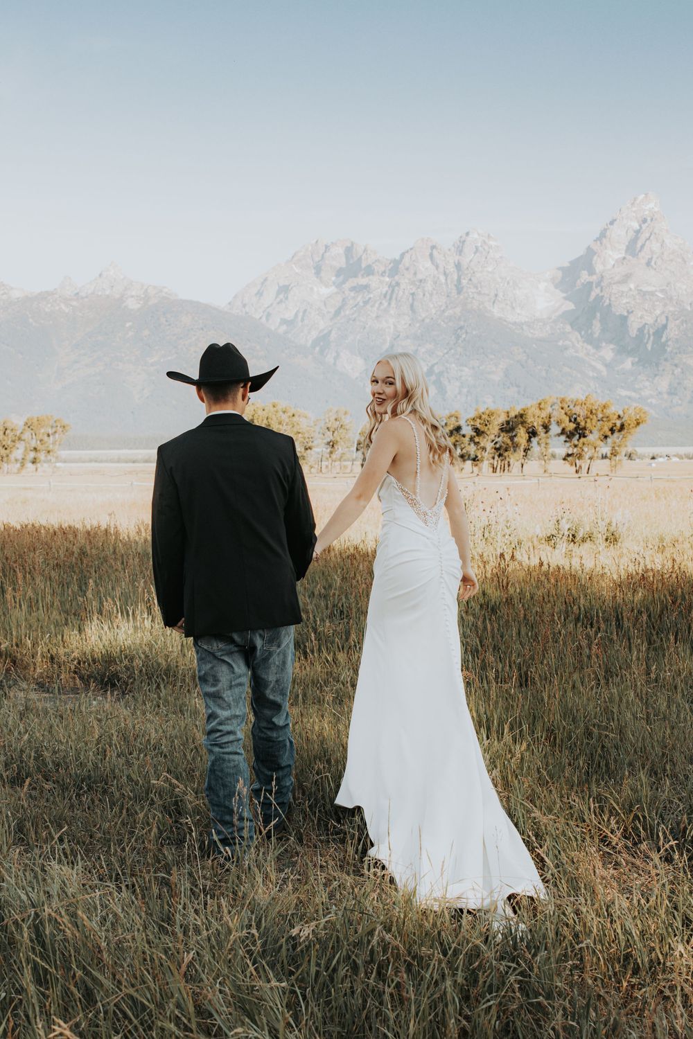 A couple walks through a rustic field with snow-capped mountains in the background at sunset.