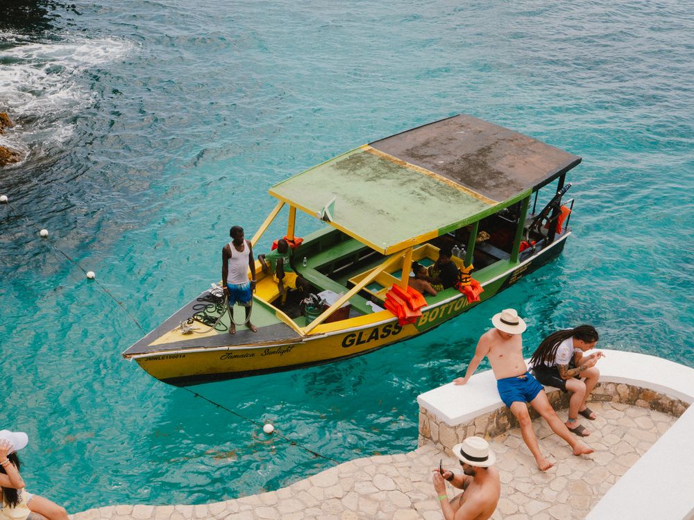 Wooden boats glide across crystal clear turquoise waters while tourists enjoy swimming from rocky shoreline in Thailand.