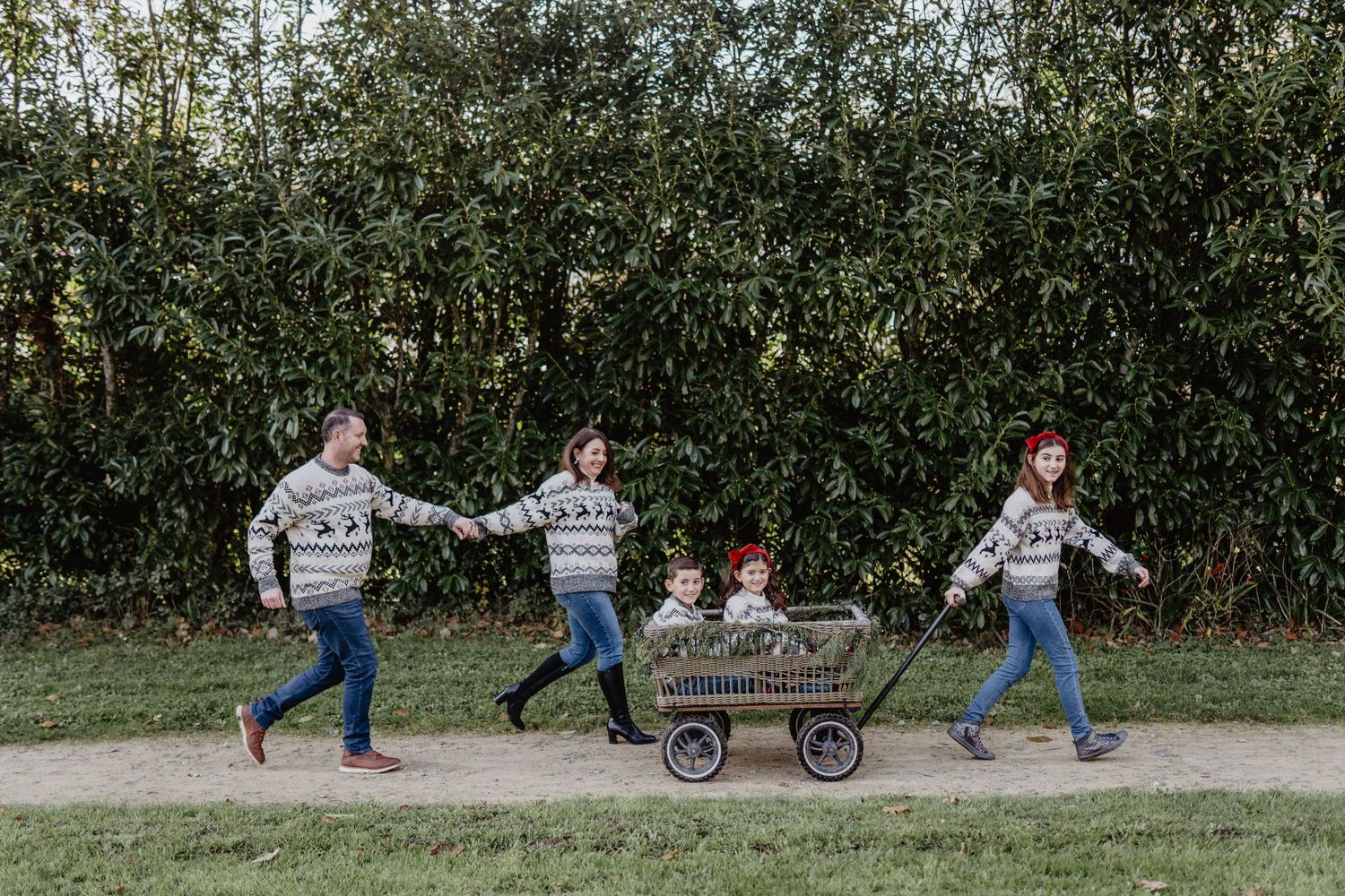 People in matching sweaters walk along a path pulling a rustic wagon through an orchard setting.