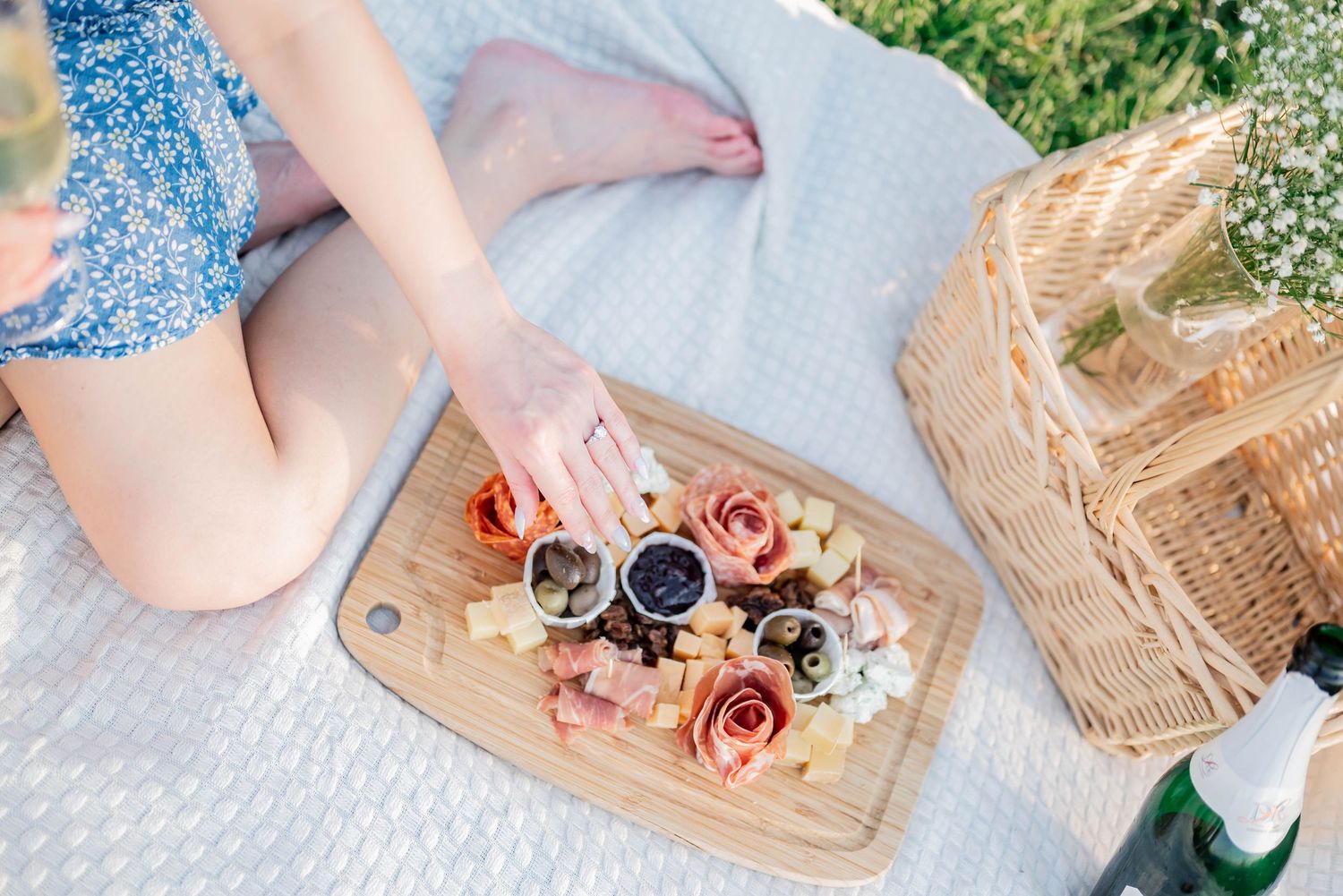 A rustic picnic setting with flowers, basket, and wine on a white blanket in a grassy outdoor location.