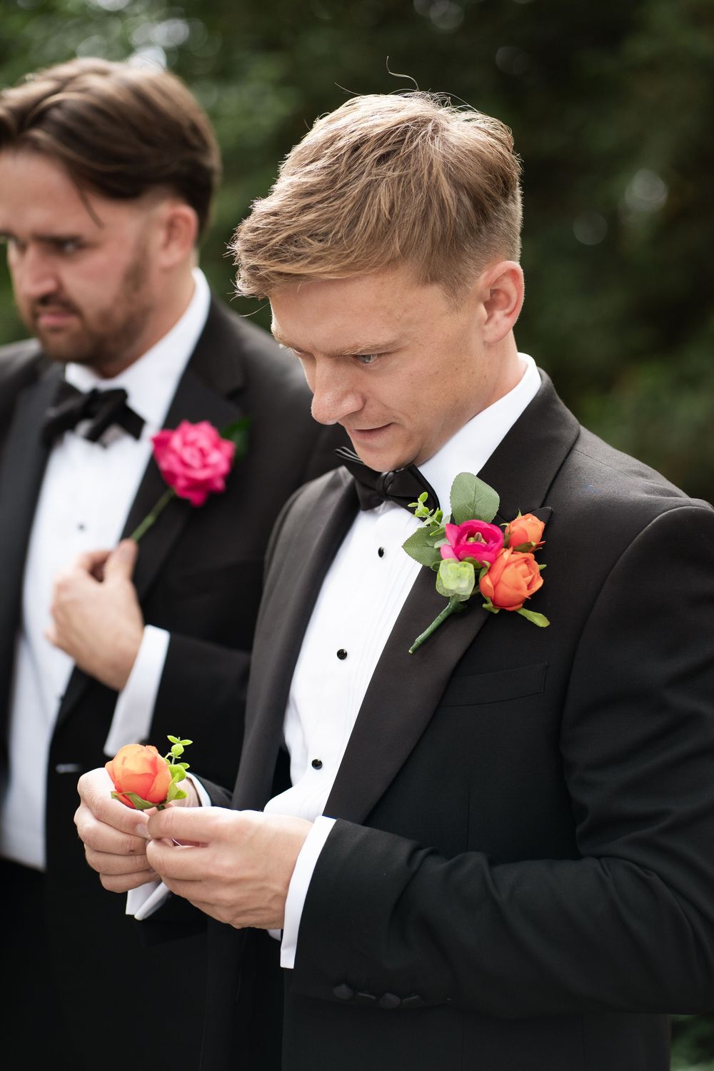 Groomsmen in black tuxedos with pink and orange boutonnieres examine their flowers at an outdoor wedding ceremony.