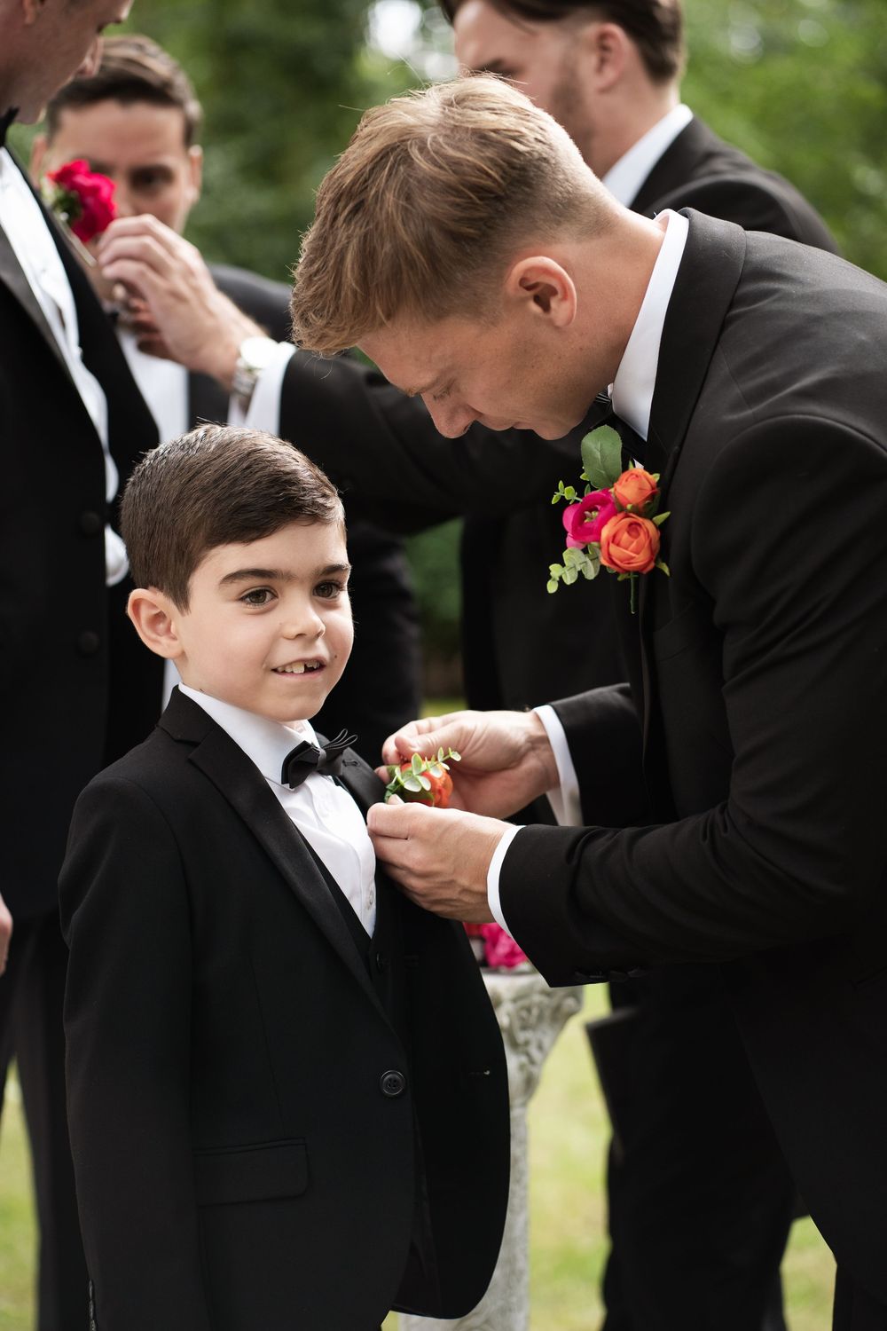 A tender moment between a man in a black suit putting on a boutonniere and a young boy at a formal occasion.