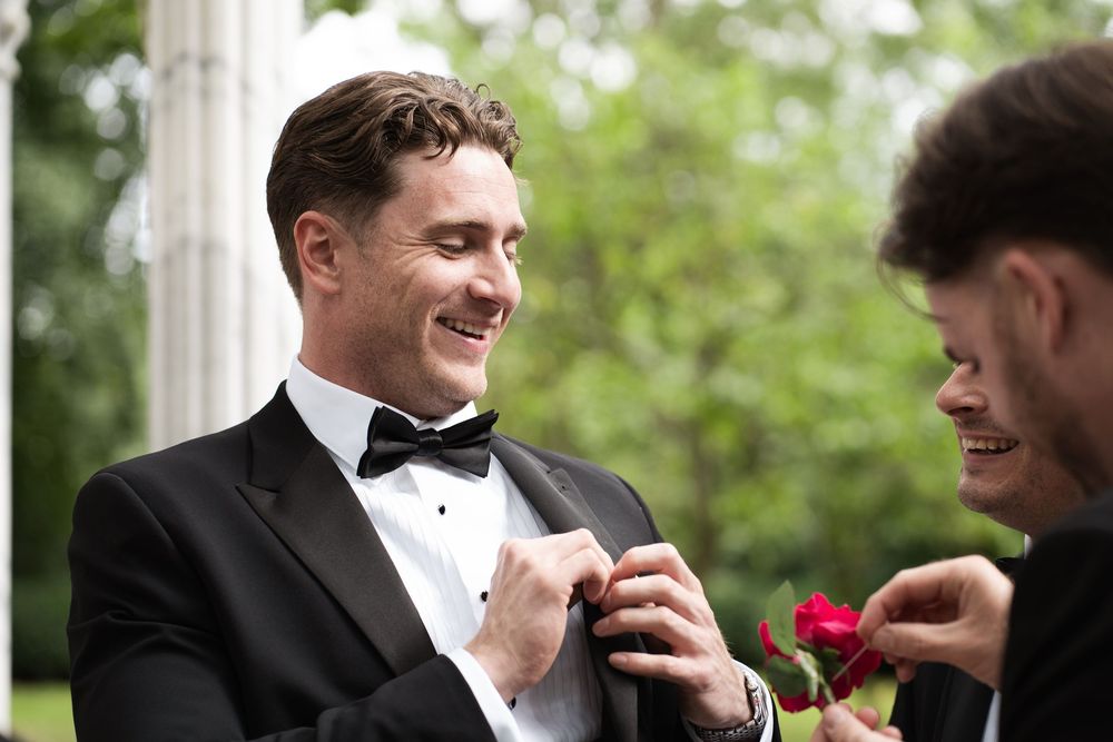 A group of groomsmen in black tuxedos share a candid moment of laughter together outdoors.