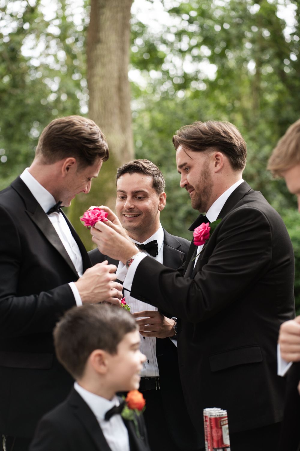 Groomsmen in black tuxedos share candid moments and laughter during a wedding celebration outdoors.