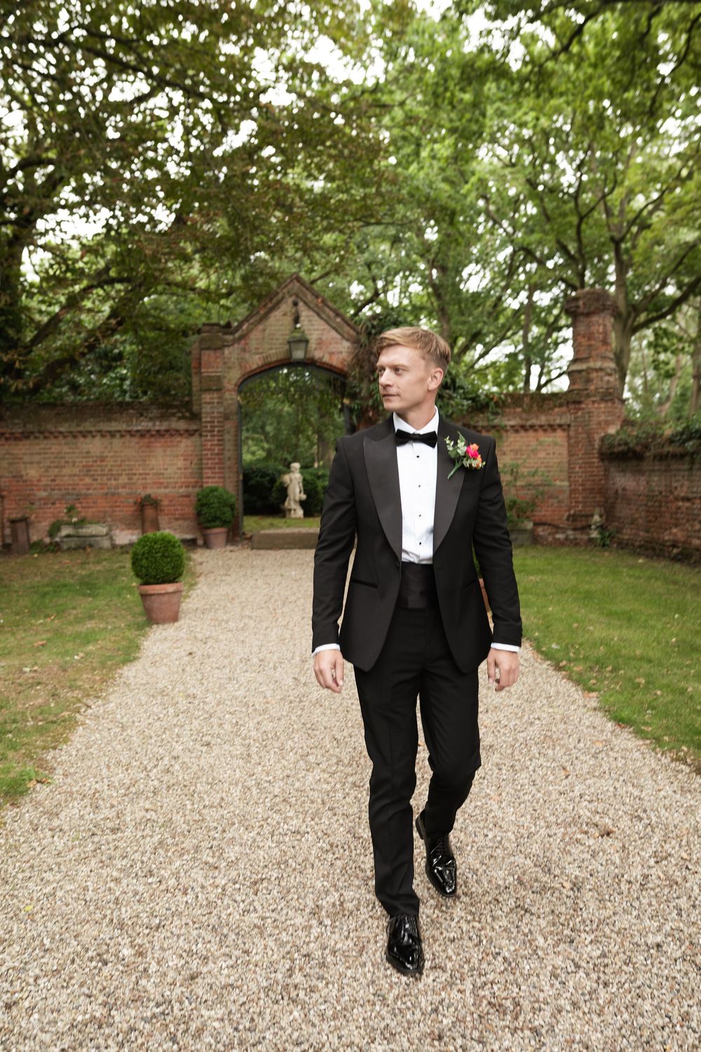 A groom in a classic black tuxedo walks along a gravel path bordered by brick walls in a garden setting.