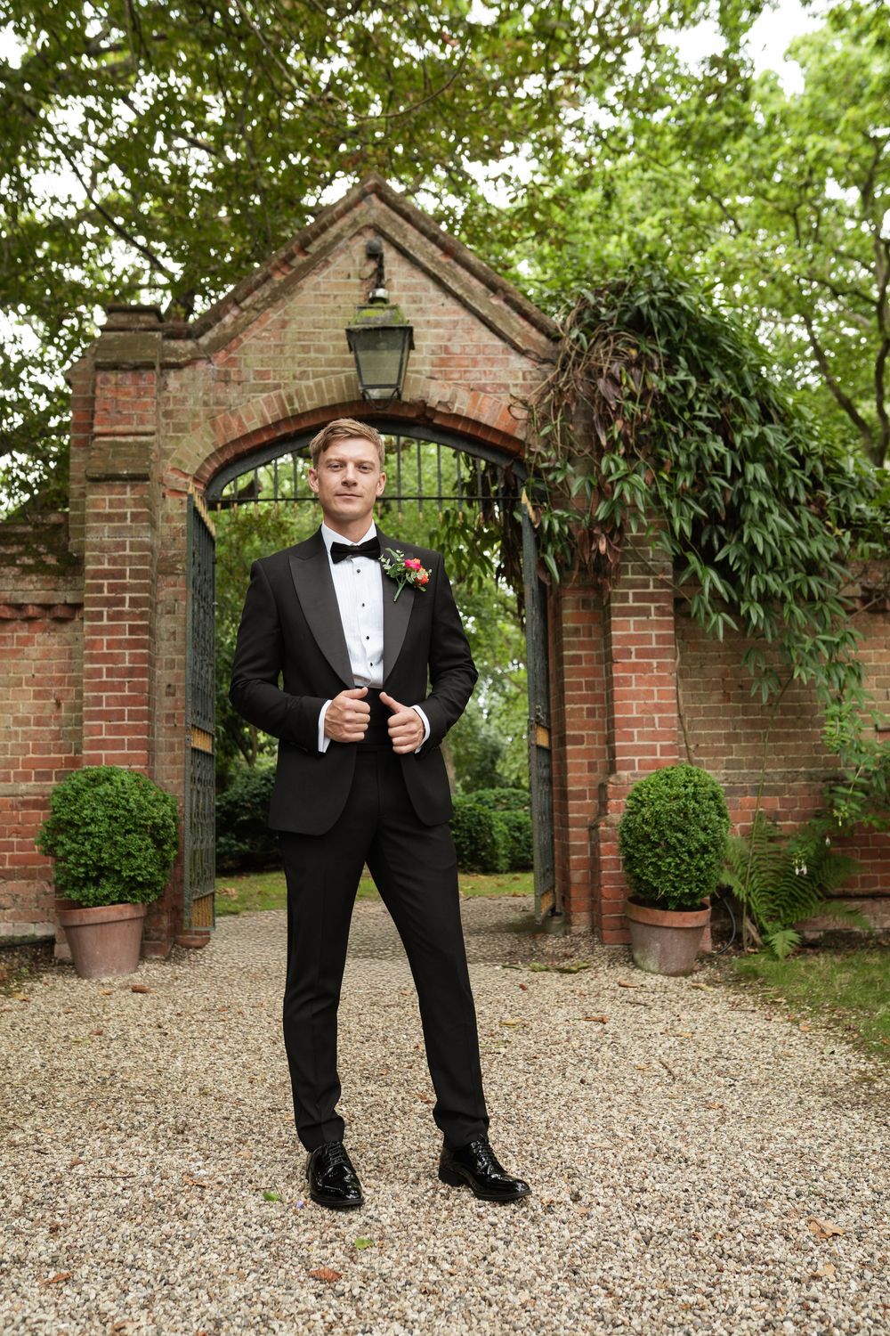A person in formal attire stands along a brick archway and gravel path in a garden setting.