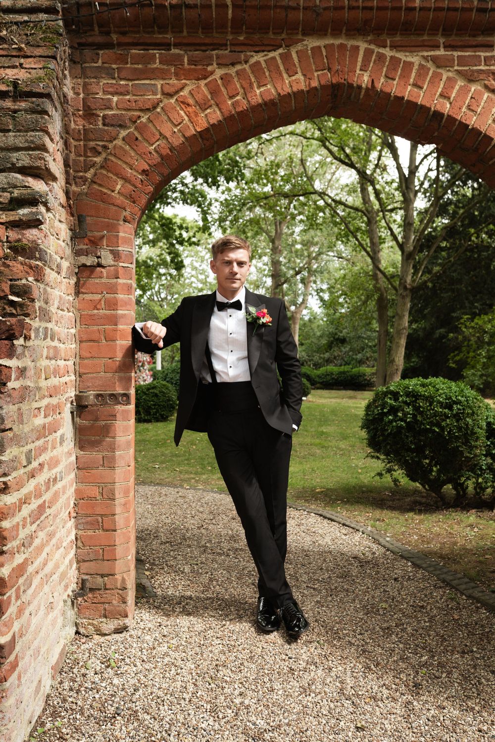 A classic black tuxedo and bow tie look photographed under a brick archway in a garden setting.