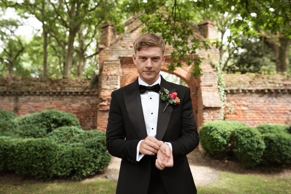 Person in formal black tuxedo adjusts bowtie while standing in lush garden setting surrounded by brick walls.
