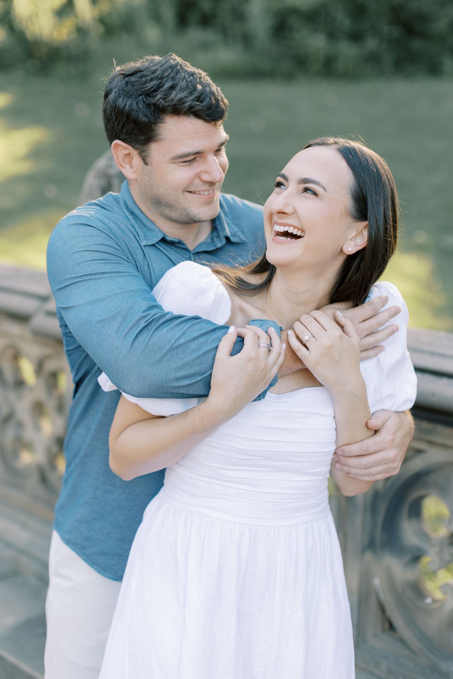 A couple in white and blue clothing share playful moments and laughter during a romantic outdoor photoshoot.