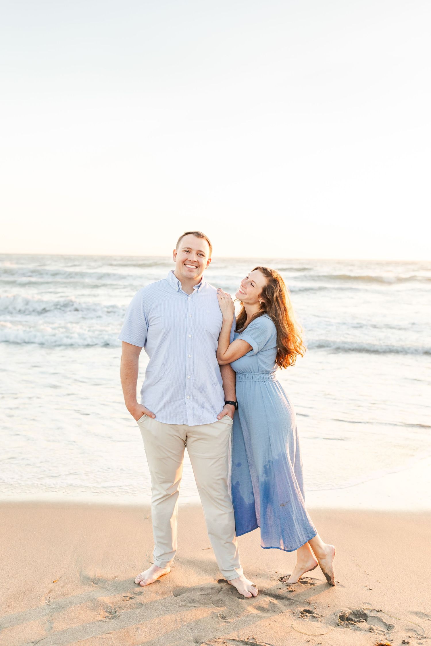 A couple in coordinated blue and white outfits poses together on a sandy beach at sunset.
