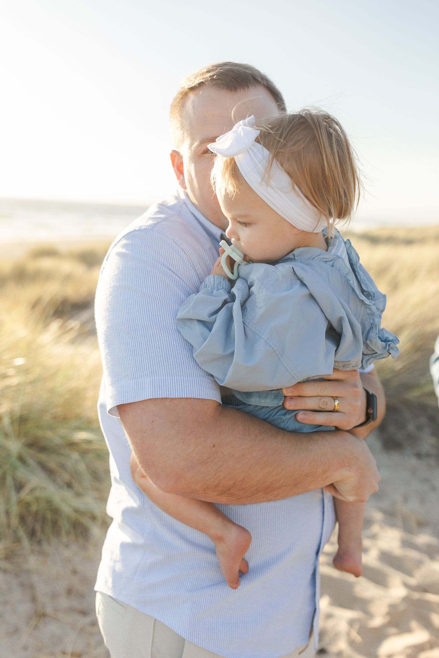 A parent in a light blue shirt holds a small child wearing denim against a beachy sand dune backdrop in warm lighting.Film by Chelsee Rawe P