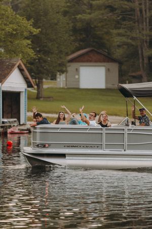 A pontoon boat filled with celebrating passengers cruises past lakeside boathouses on a summer day.