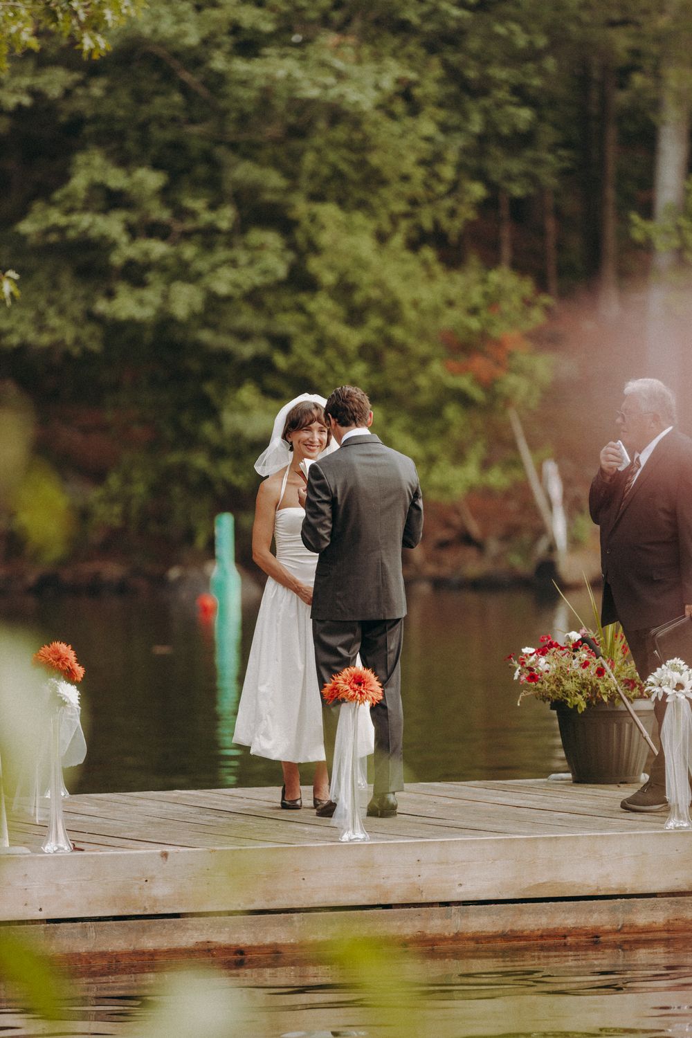 A ceremony takes place on a wooden dock decorated with orange flowers beside a serene lake.