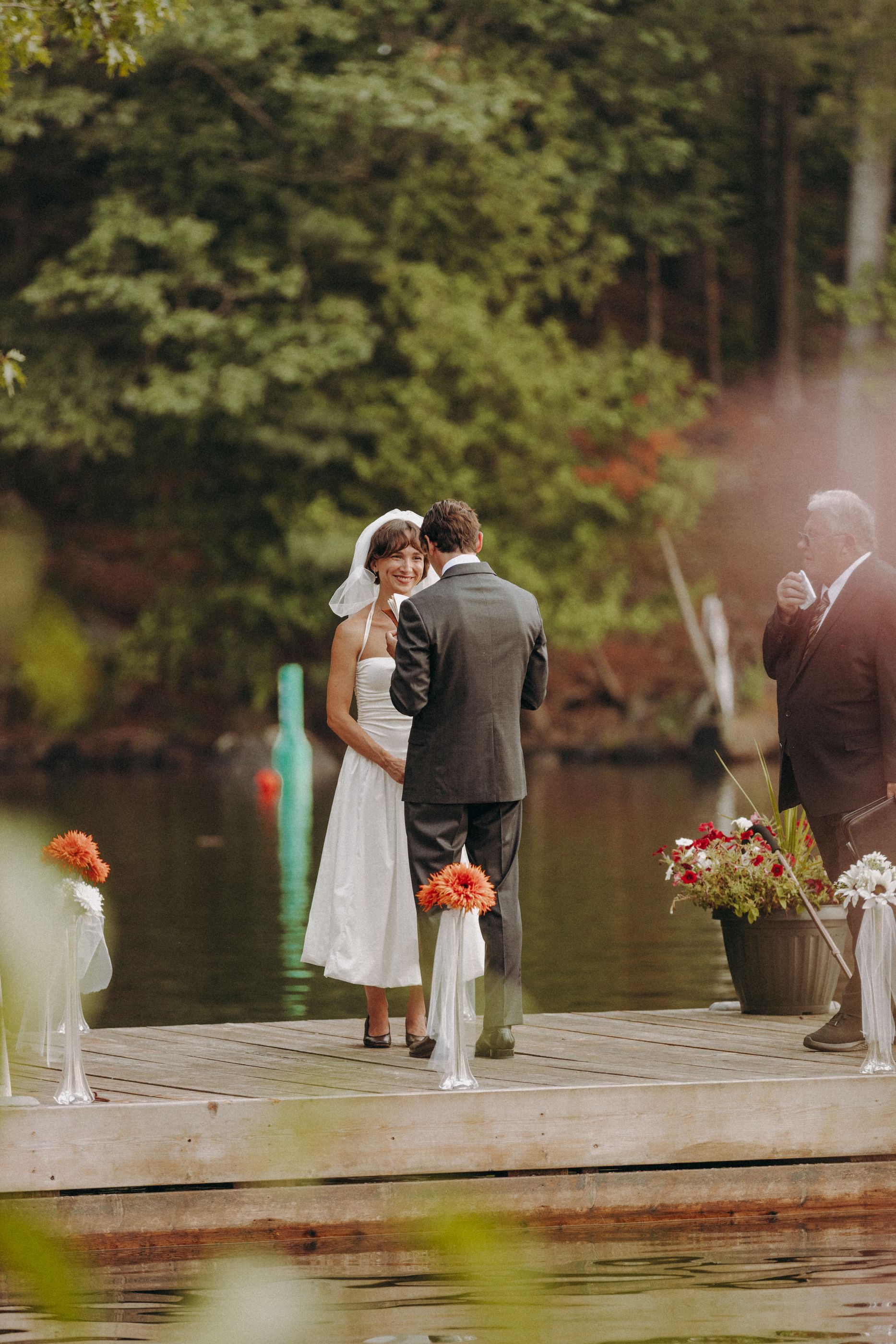 A ceremony takes place on a wooden dock decorated with orange flowers beside a serene lake.