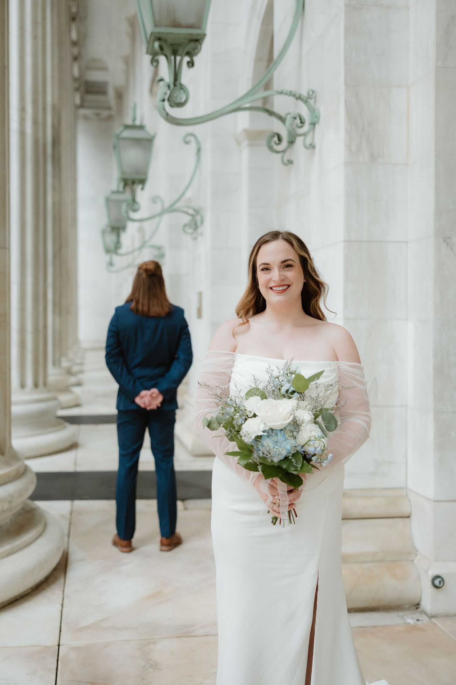 Wedding party members in formal attire stand in an elegant white columned venue with decorative green fixtures.