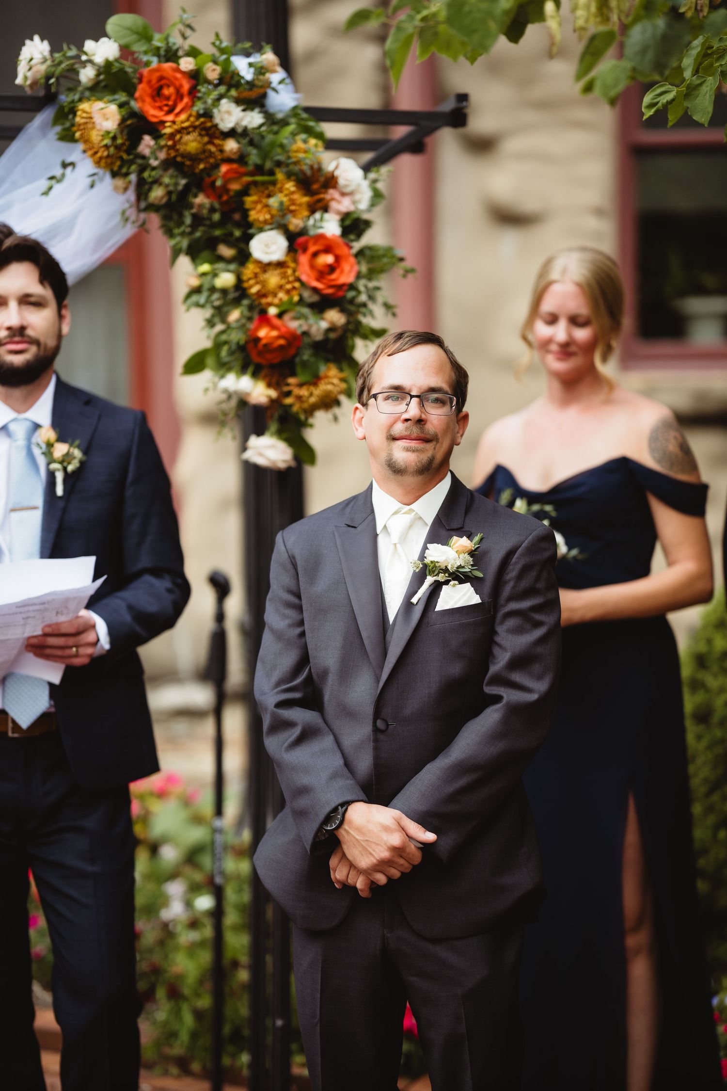 Wedding party stands under floral arch decorated with orange roses and greenery against brick building exterior.