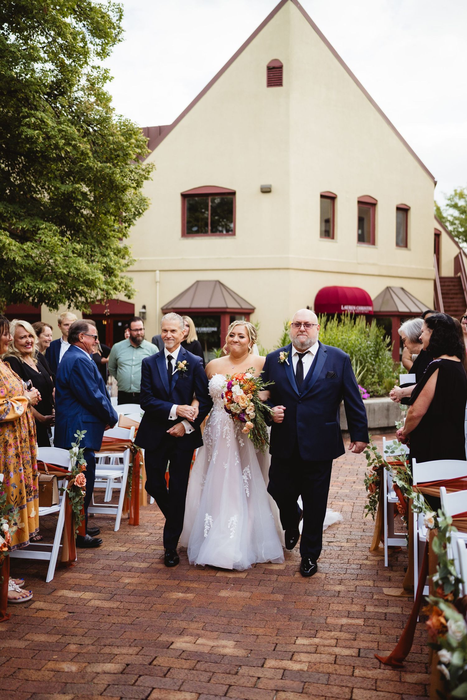 A wedding ceremony procession takes place on a brick pathway outside a white church building under green trees.