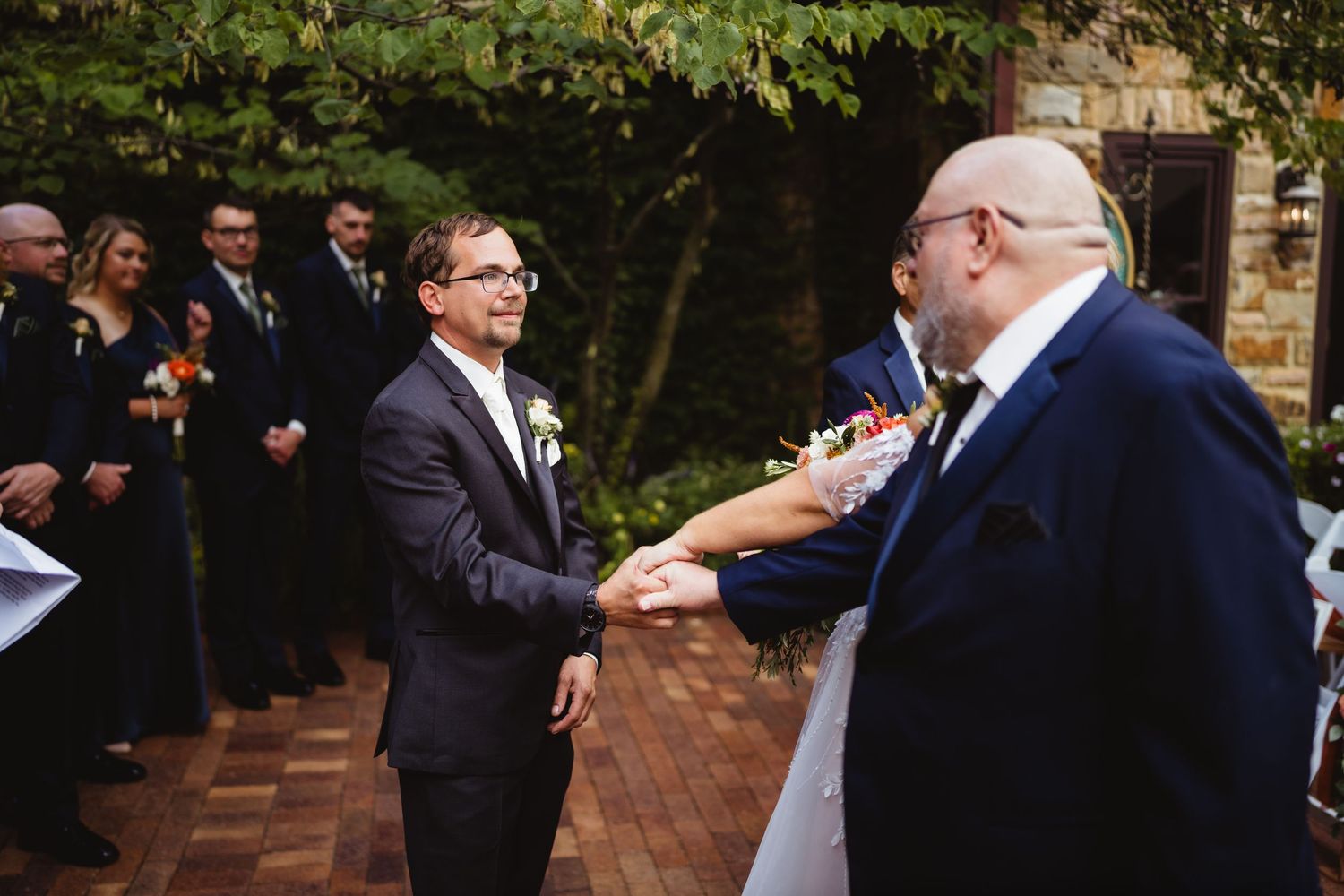 Two people share an emotional moment during an outdoor wedding ceremony on a brick patio with guests looking on.